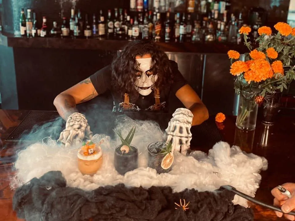 Persona con maquillaje de calavera presenta bebidas entre niebla y flores de cempasúchil en una barra.