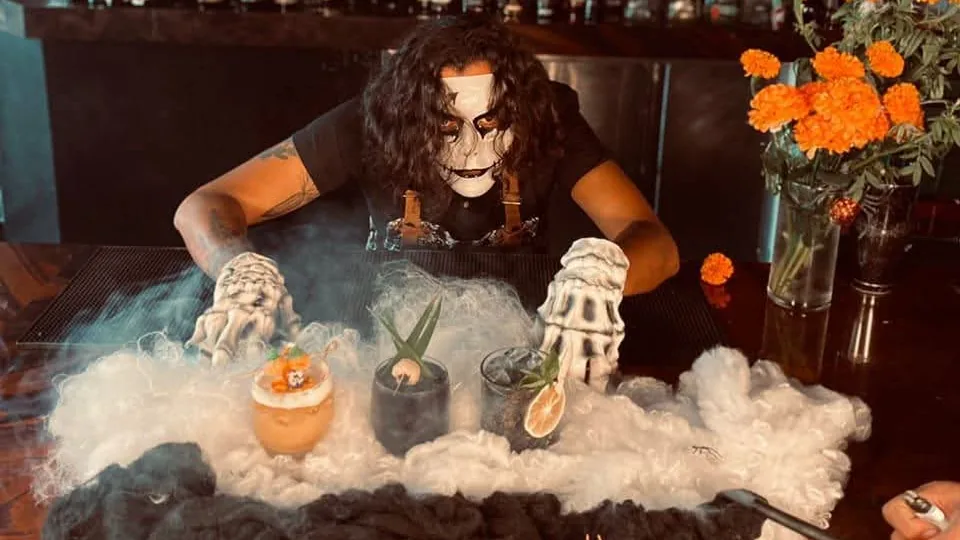 Persona con maquillaje de calavera presenta bebidas entre niebla y flores de cempasúchil en una barra.