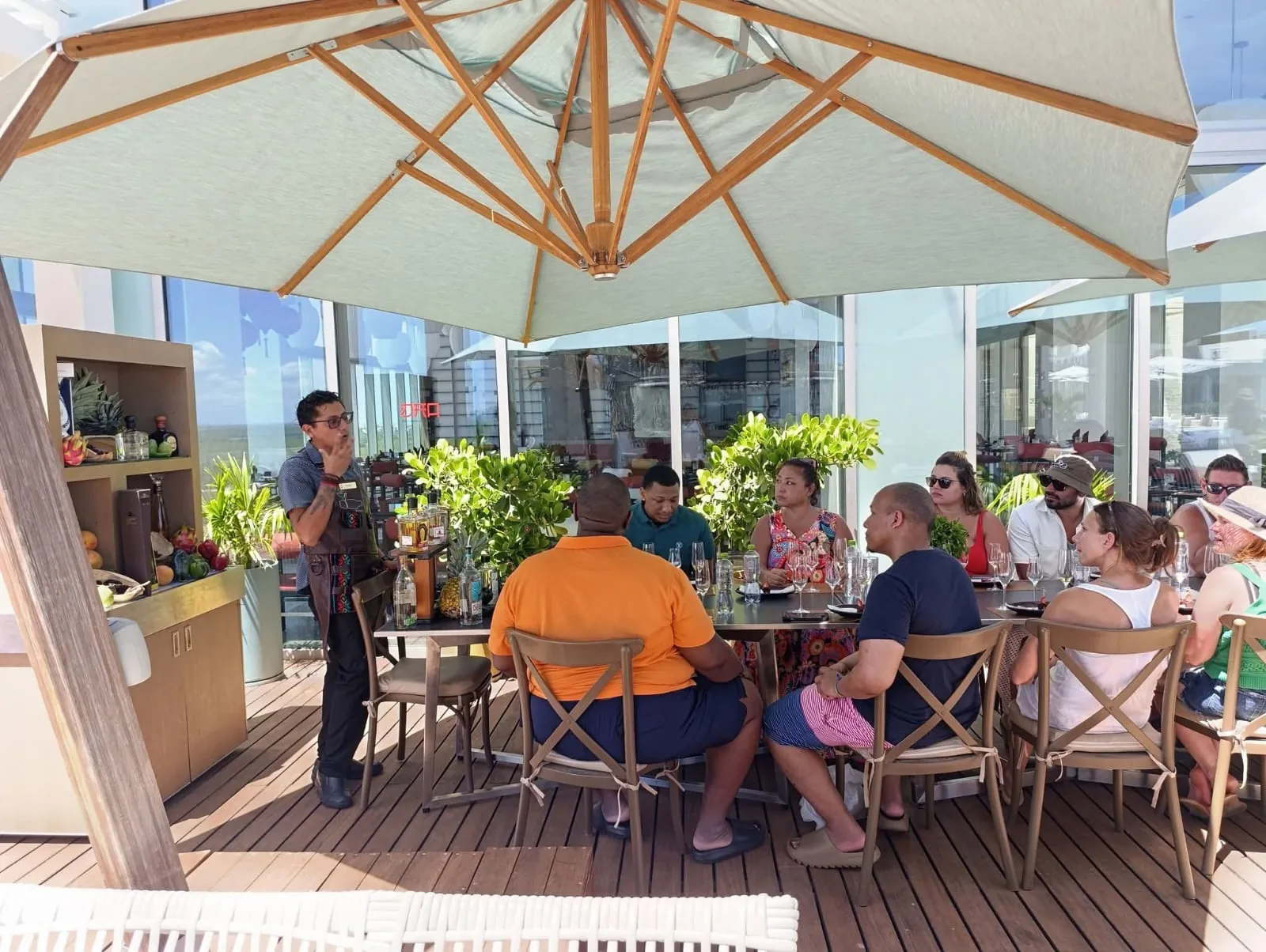 Una persona con mandil explica una degustación a un grupo sentado en una terraza bajo una sombrilla.